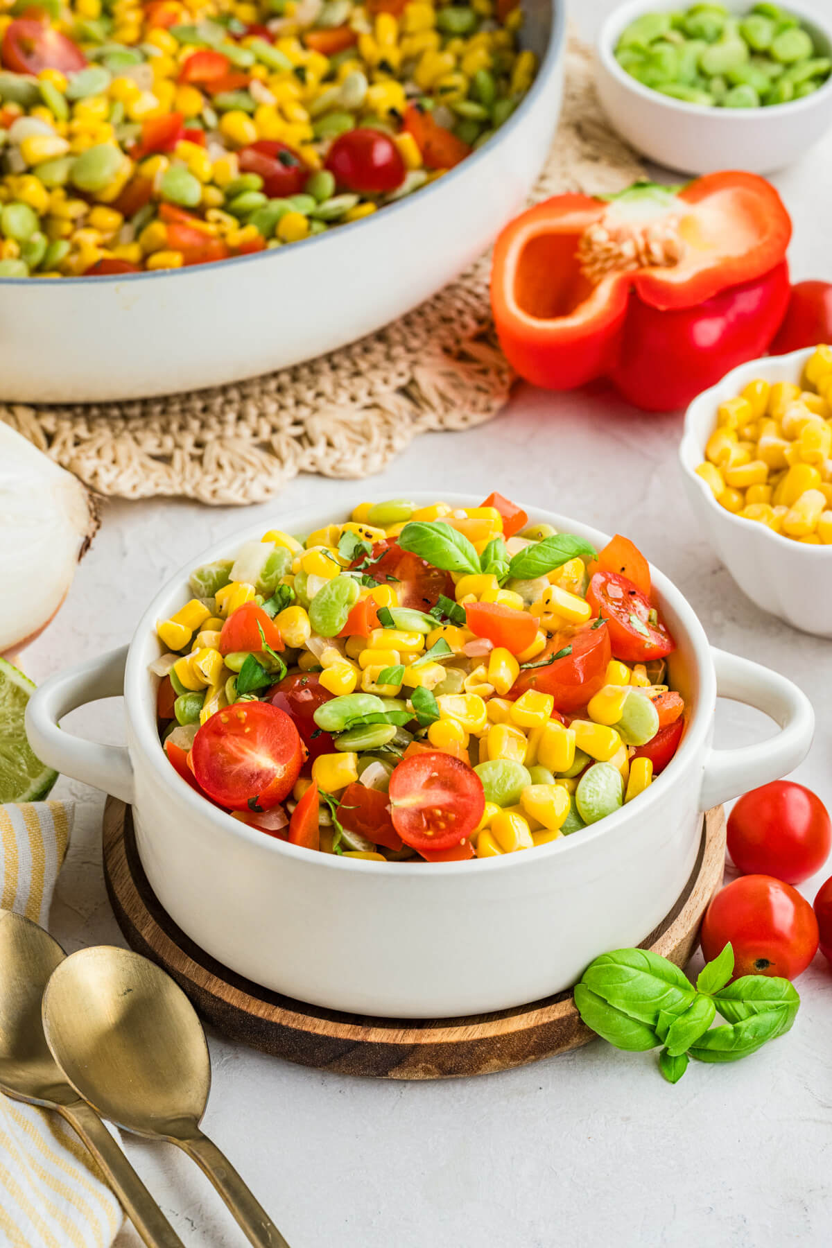 A white serving dish filled with vibrant yellow corn succotash featuring green lima beans, red bell peppers, cherry tomatoes, and fresh basil on a dinner table.