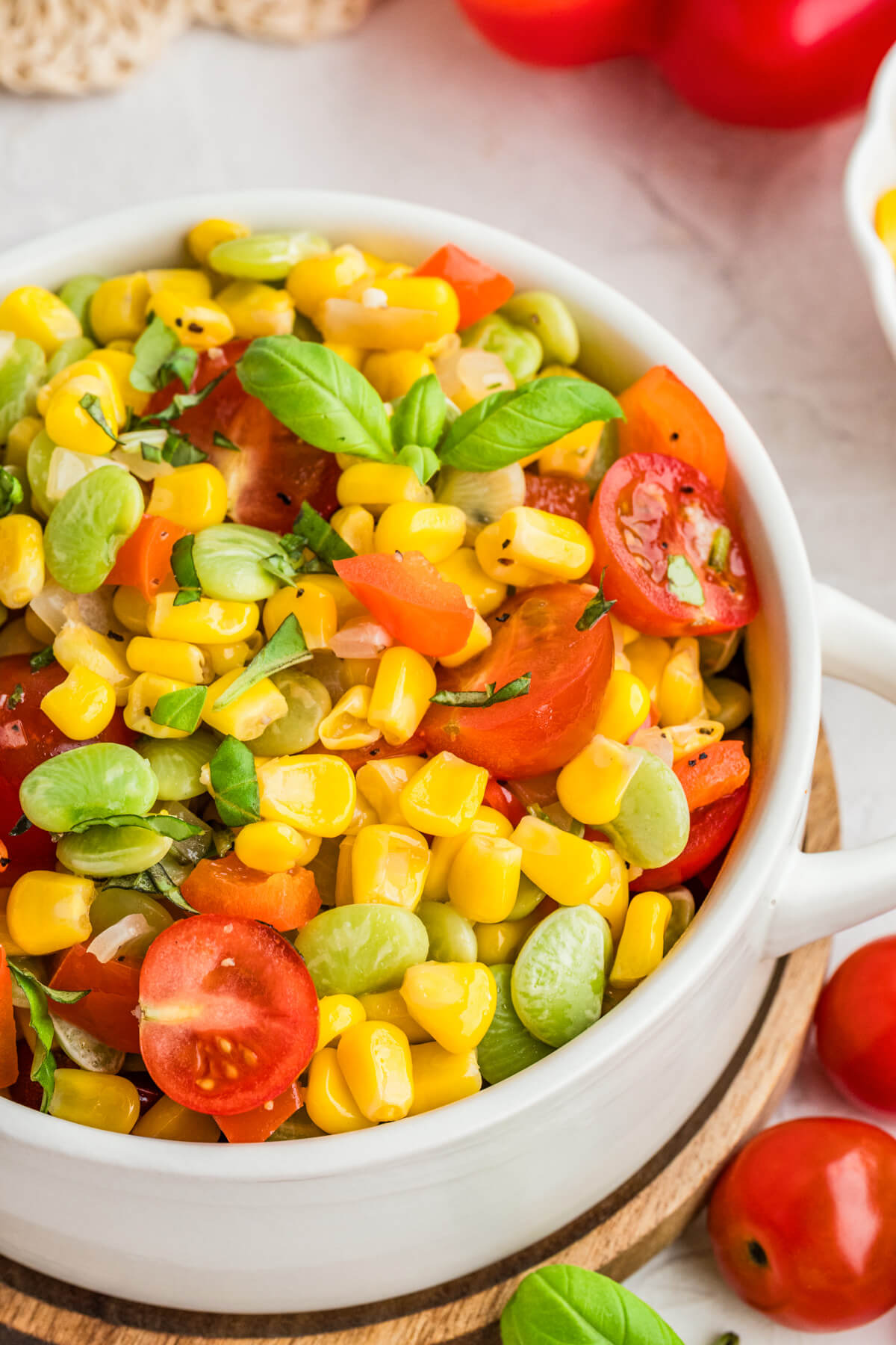 A white serving dish filled with vibrant yellow corn succotash featuring green lima beans, red bell peppers, cherry tomatoes, and fresh basil.