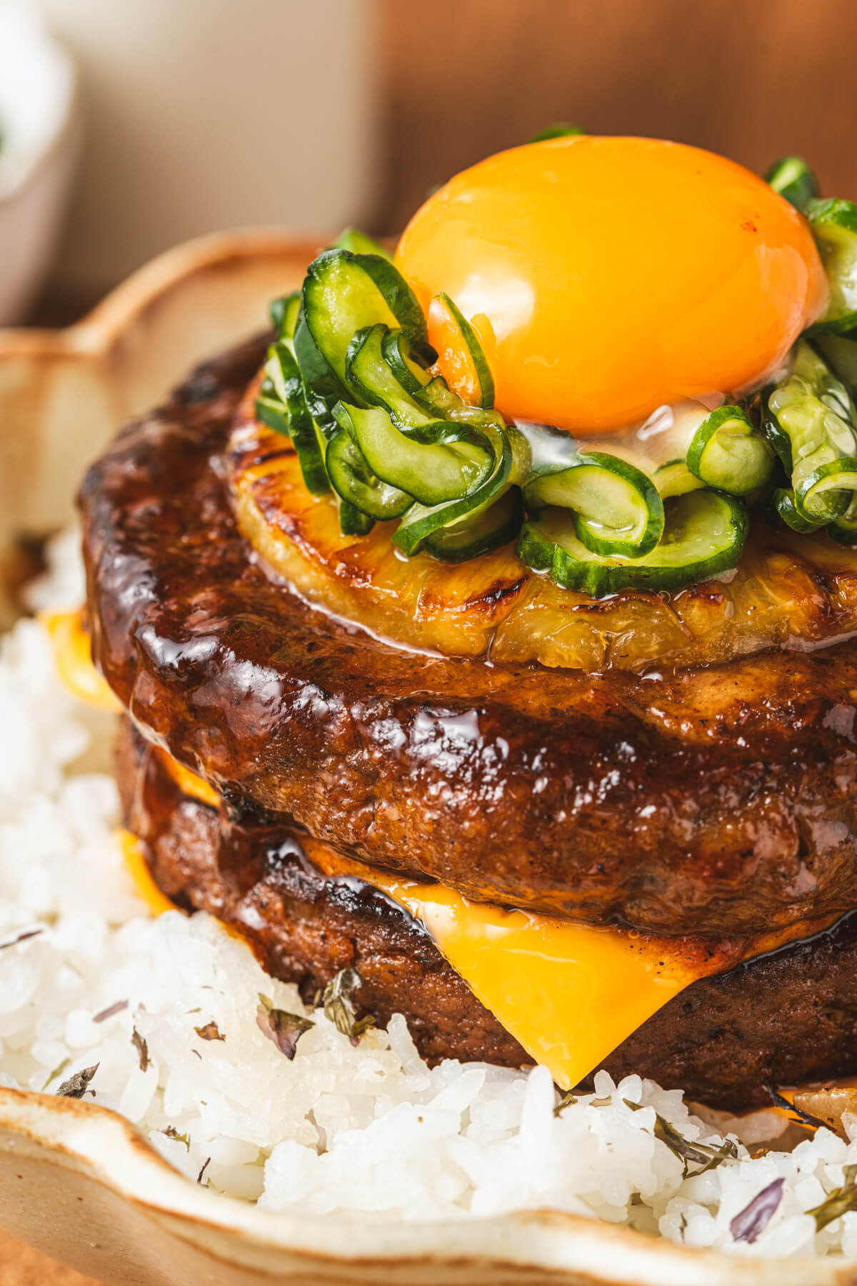 A two layer Japanese Hambagu steak dish piled high with American cheese, grilled pineapple, picked cucumber and topped with an egg yolk on a bed of seasoned rice.