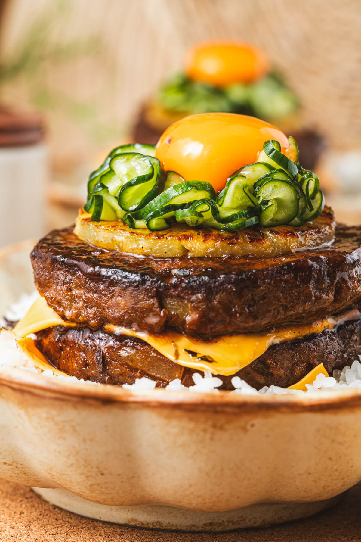 A two layer Japanese Hambagu steak dish piled high with American cheese, grilled pineapple, picked cucumber and topped with an egg yolk on a bed of seasoned rice.