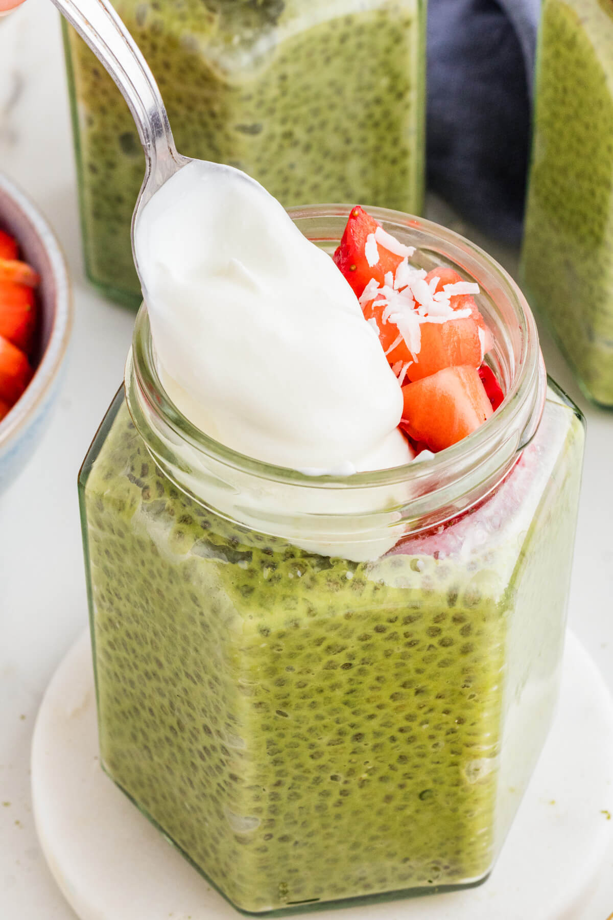 A spoon drops a dollop of coconut cream onto the top of a jar of coconut matcha chia pudding.