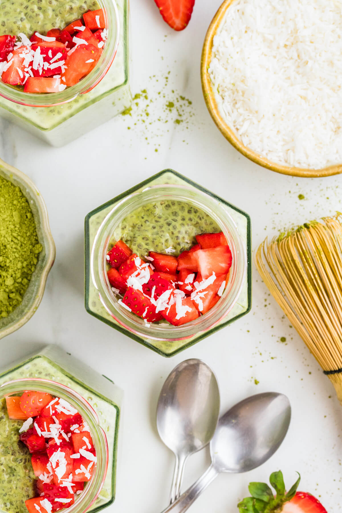 Overhead view of hexagonal matcha chia pudding jars with matcha powder, a bamboo whisk, spoons, and coconut cream.