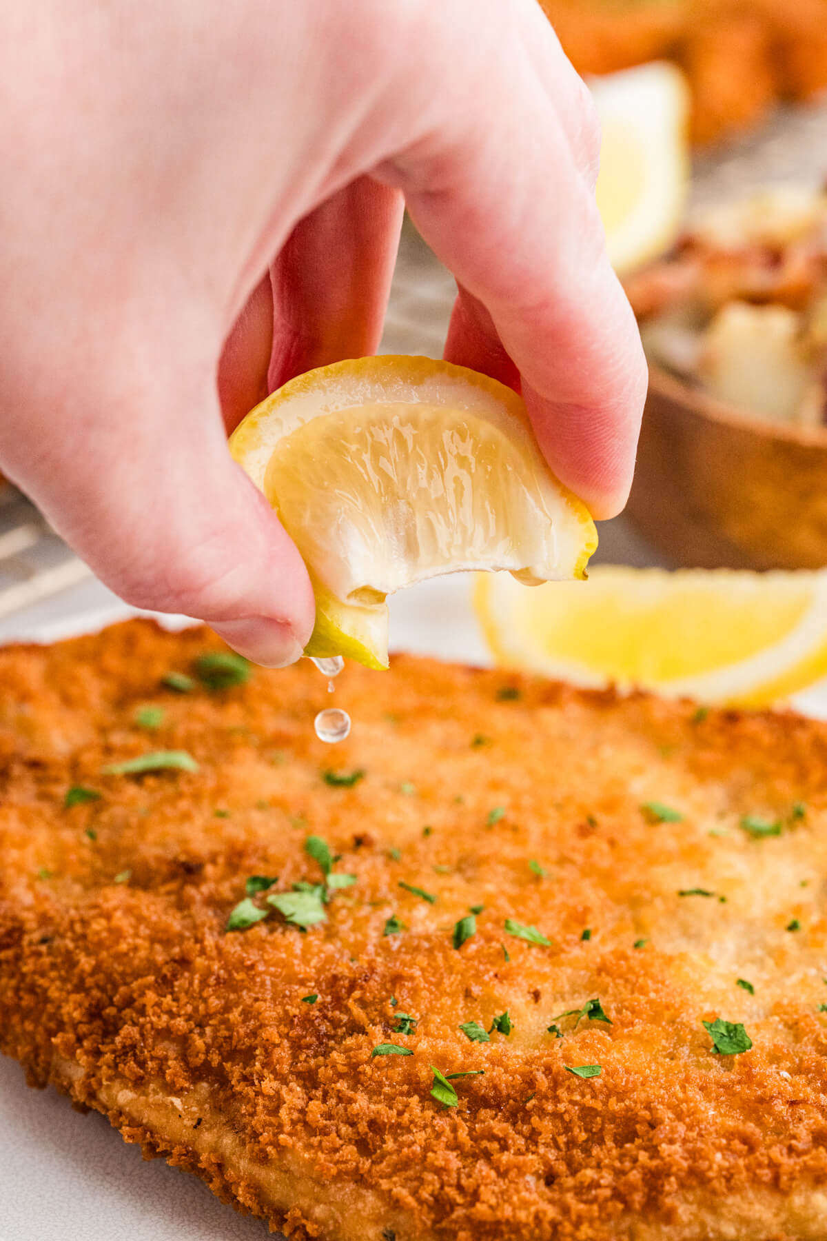 A hand drizzles juice from a lemon wedge over a pork schnitzel.