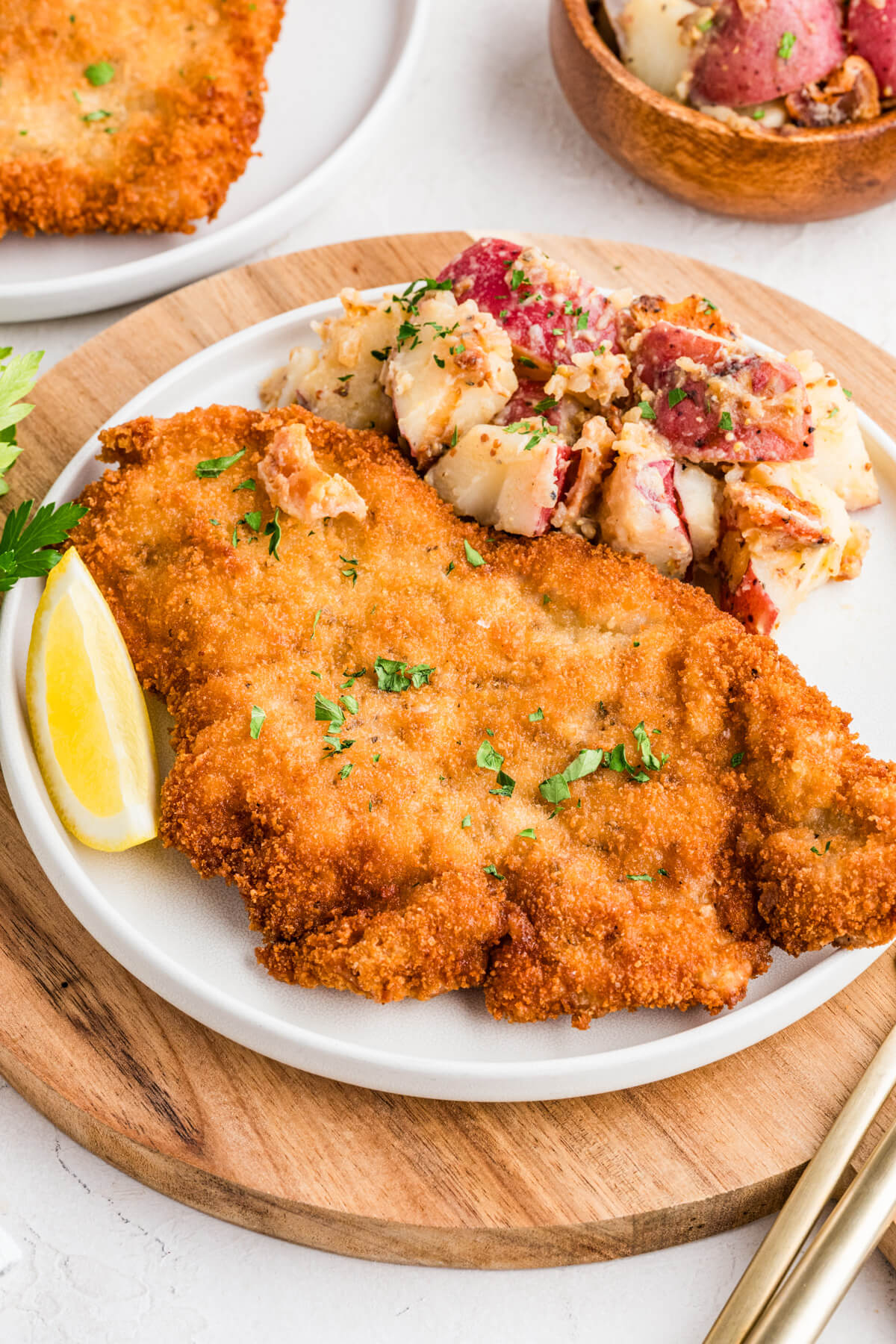 A golden brown fried pork schnitzel on a plate with a lemon wedge and a side of potato salad.