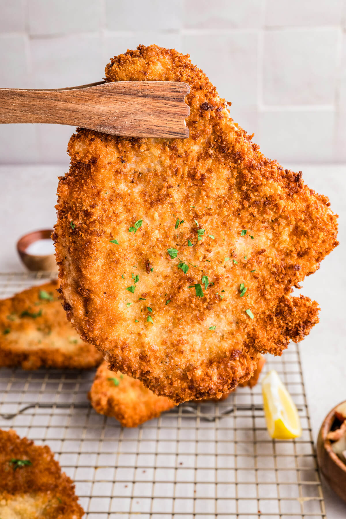 A pair of wooden tongs holds a golden fried pork schnitzel in the air.