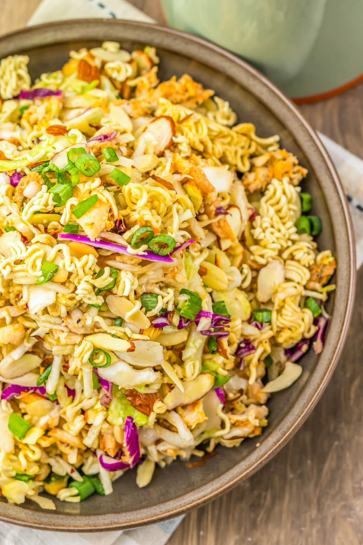 A brown bowl filled with ramen noodle salad featuring pulled rotisserie chicken, coleslaw mix, sesame seeds, and green onions.