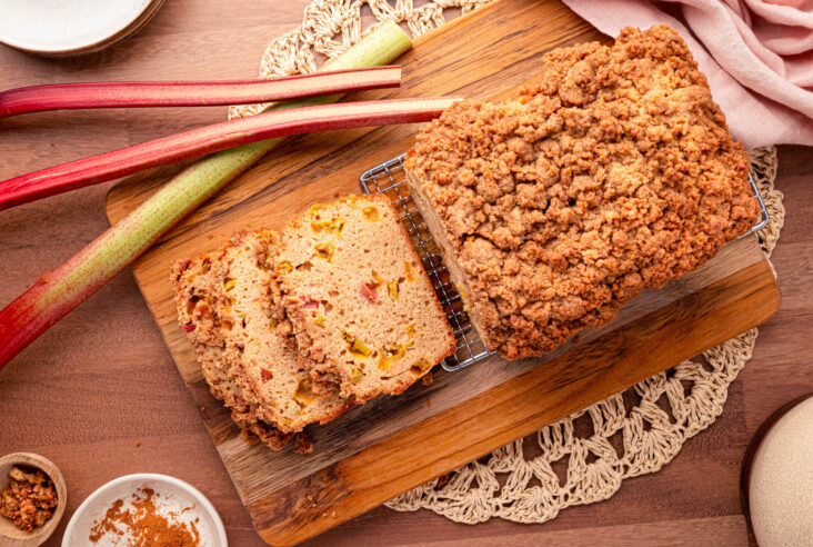 Overhead view of a partially sliced rhubarb bread on a wooden cutting board surrounded by stalks of rhubarb.