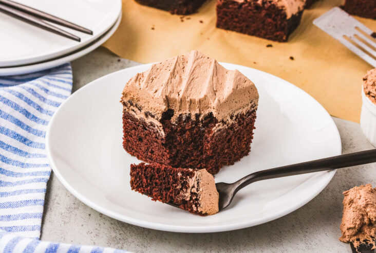 A white dessert plate holding a piece of frosted chocolate wacky cake with a bite missing.