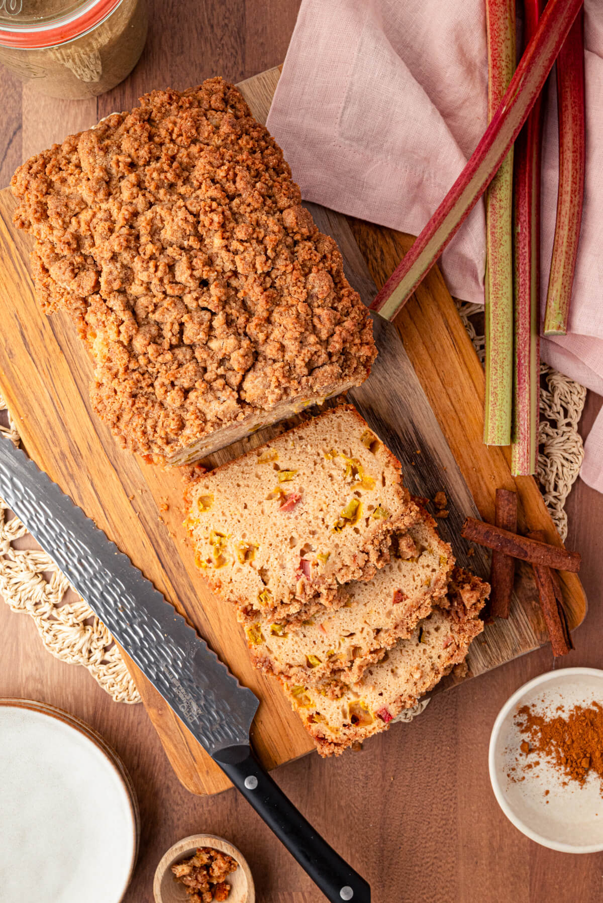 Overhead view of a partially sliced rhubarb bread on a wooden cutting board surrounded by stalks of rhubarb.