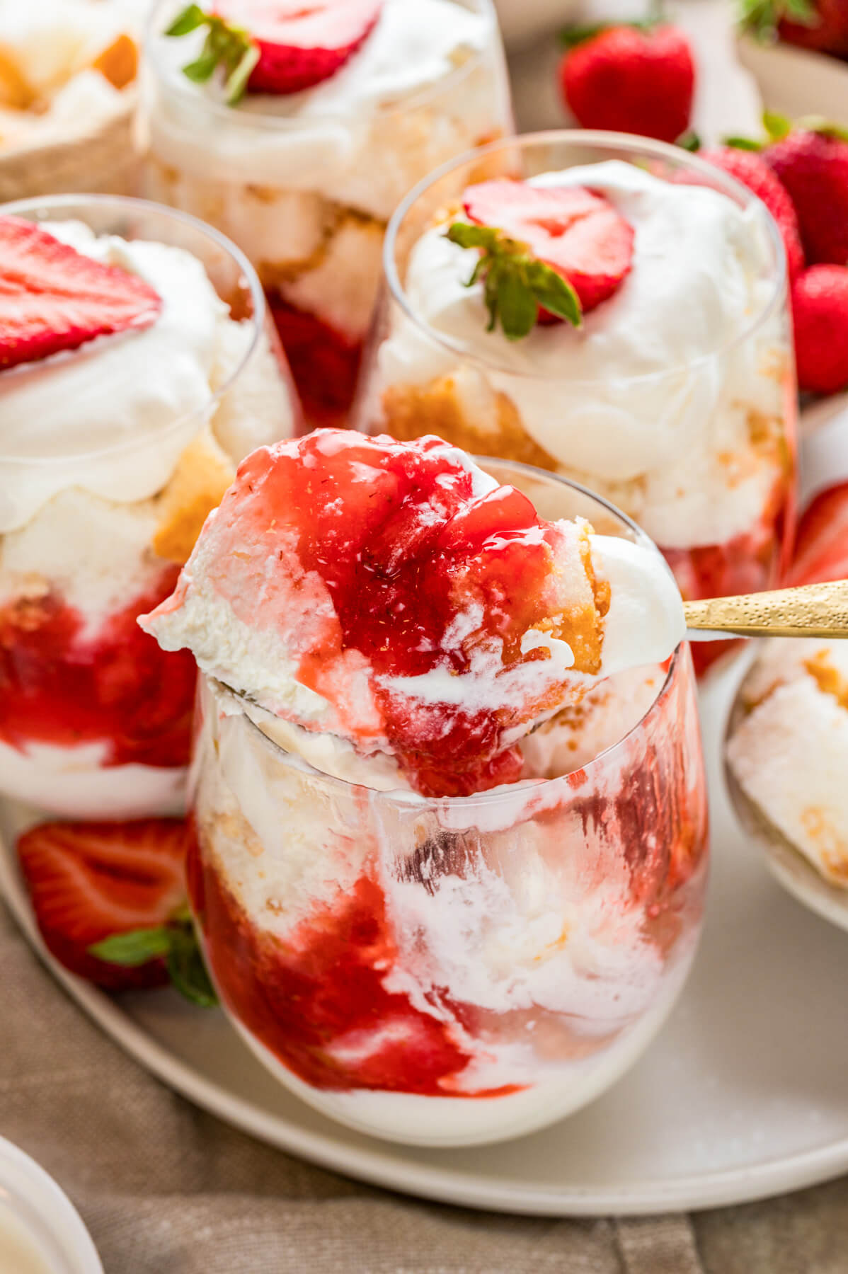A spoon holding a portion of strawberry trifle above a stemless wine glass of the same dessert.