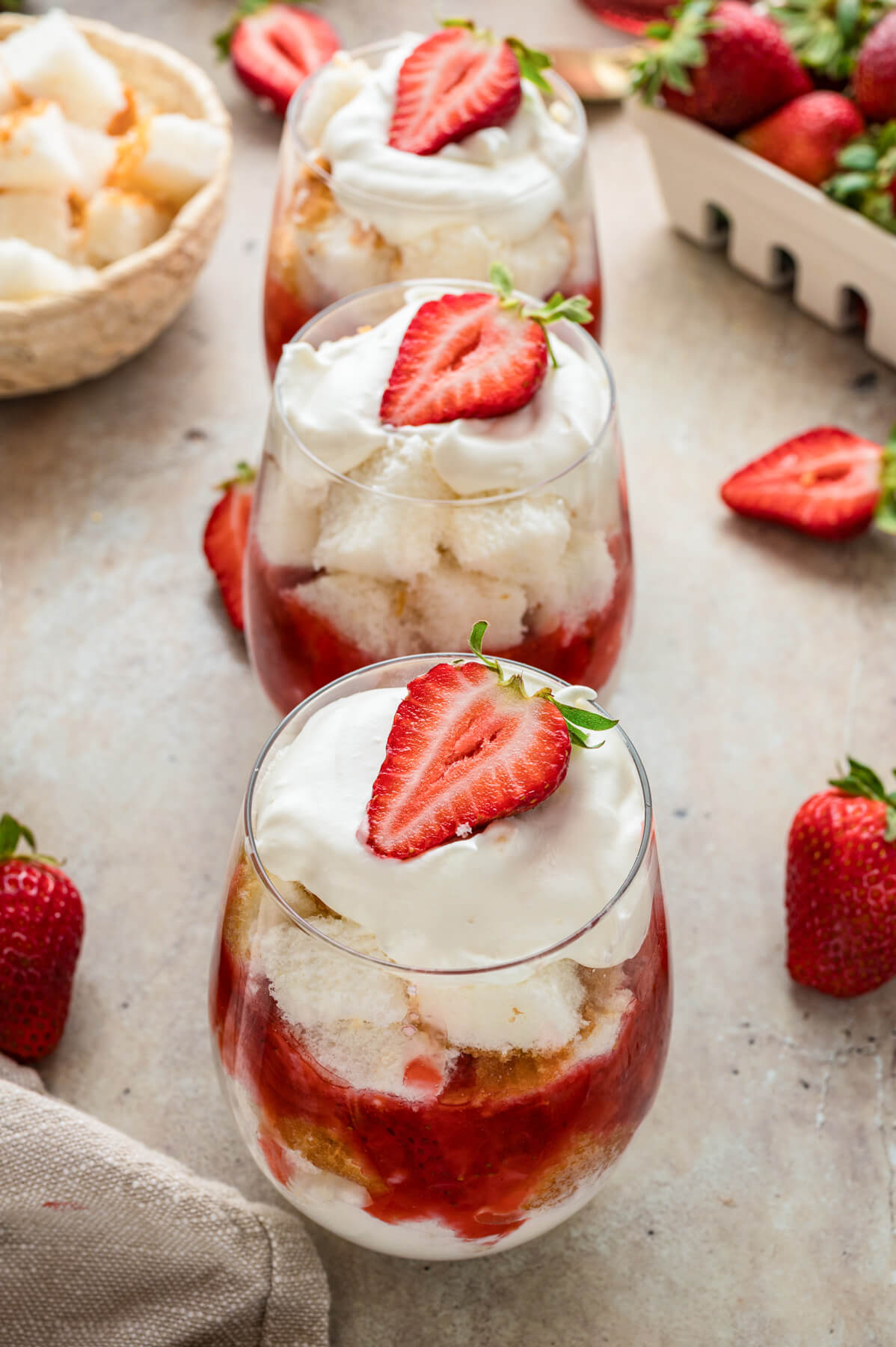 Three dreamy strawberry trifles topped with whipped cream and a sliced strawberry sit in a line on a dinner table.