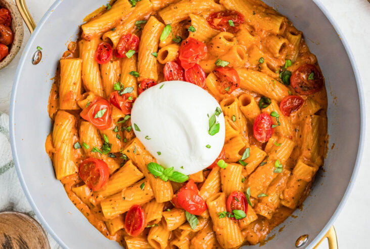 A fresh burrata ball sits on top of a pot full of pasta in roasted tomato sauce.