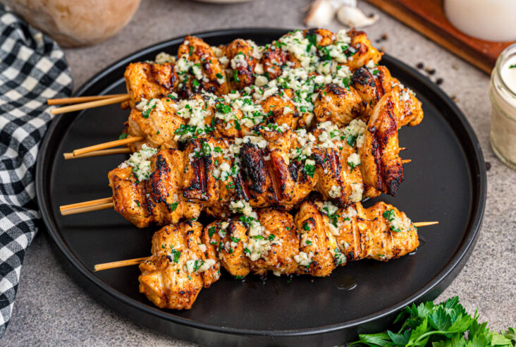 A black plate containing several perfectly grilled Garlic Parmesan Chicken Skewers loaded with parsley and garlic.