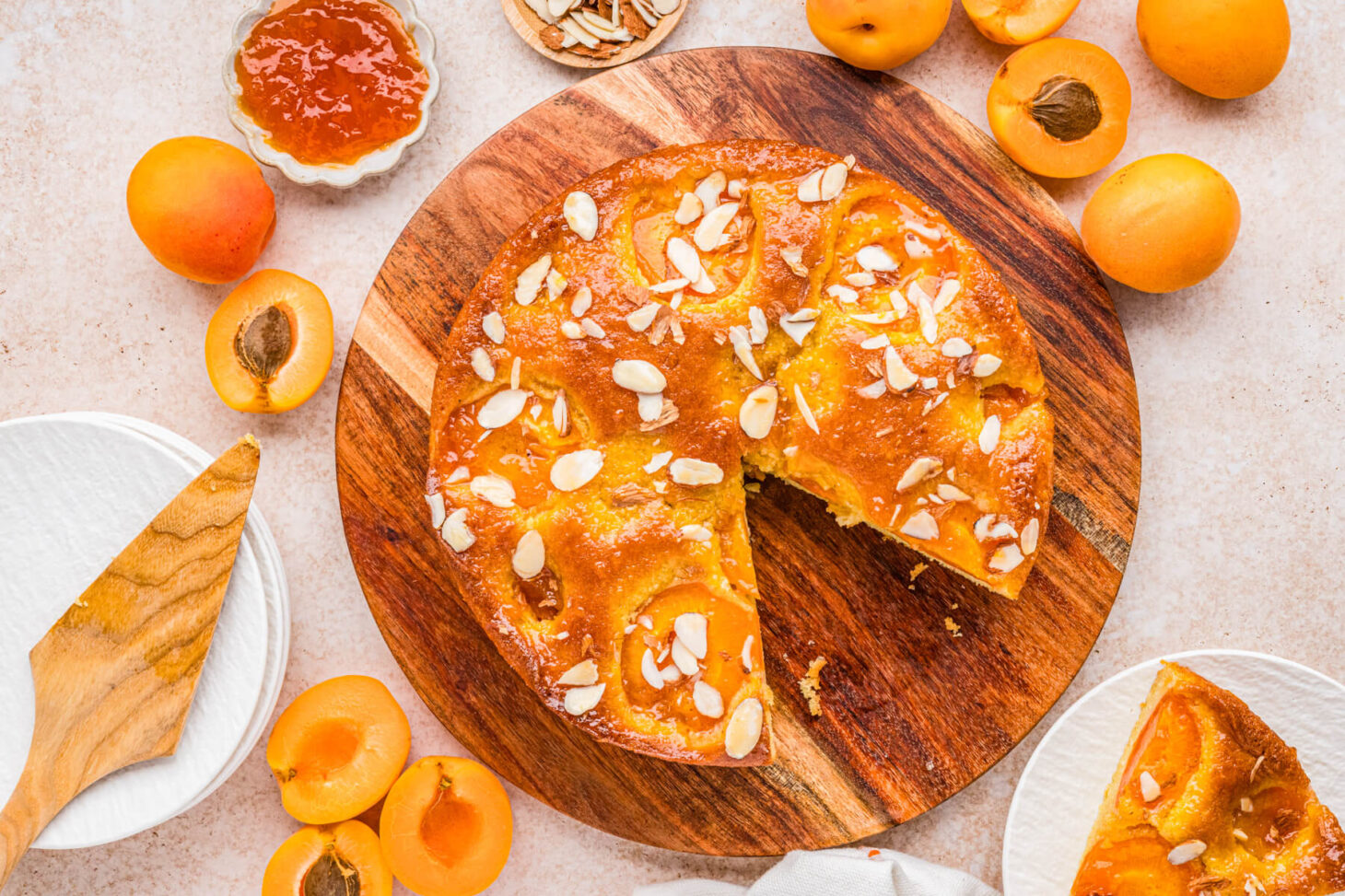 Overhead view of an almond apricot cake with one slice missing on a wooden serving board surrounded by fresh sliced apricots.