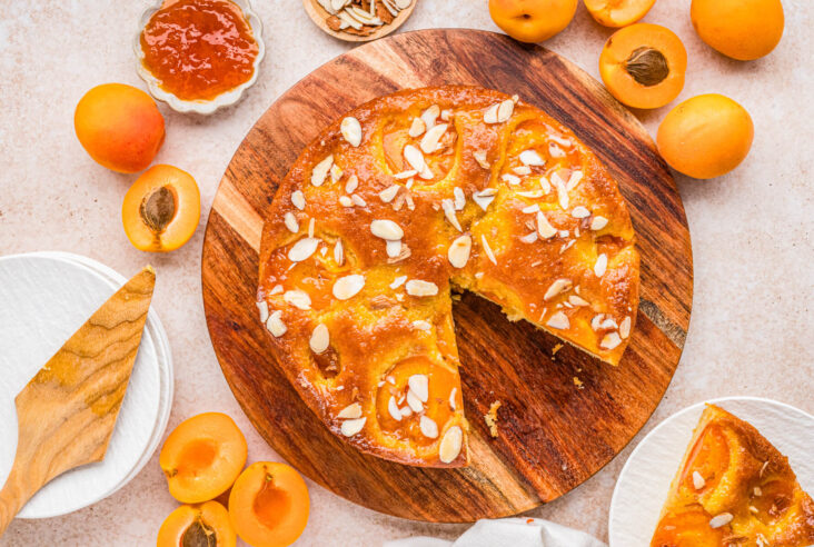Overhead view of an almond apricot cake with one slice missing on a wooden serving board surrounded by fresh sliced apricots.