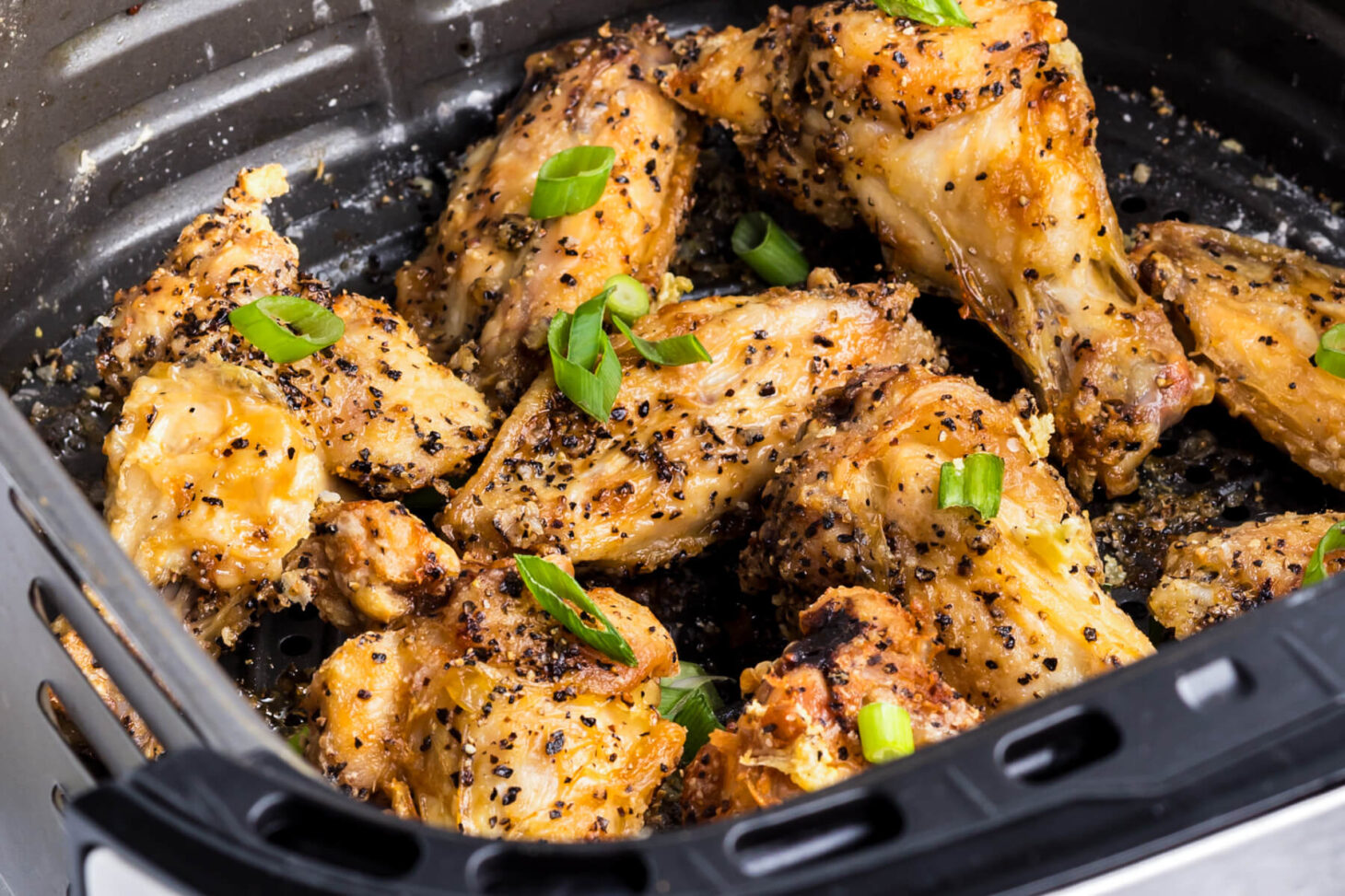 A black air fryer basket full of golden brown crispy Salt and Pepper Wings garnished with sliced green onions.