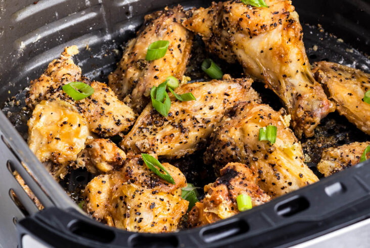 A black air fryer basket full of golden brown crispy Salt and Pepper Wings garnished with sliced green onions.