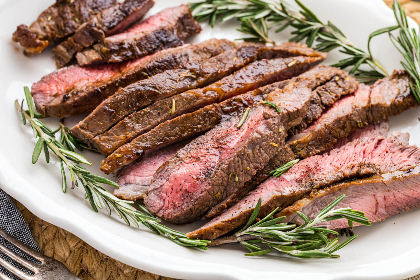 Strips of perfectly seared cast iron skillet steak surrounded by fresh rosemary on a white platter.
