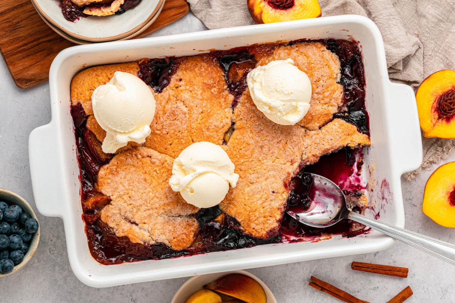A white baking dish of Peach Blueberry cobbler topped with scoops of vanilla ice cream.