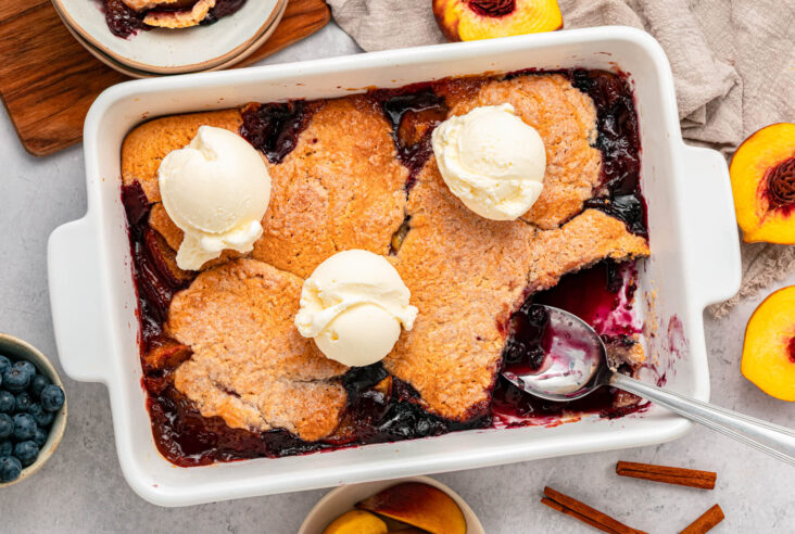A white baking dish of Peach Blueberry cobbler topped with scoops of vanilla ice cream.