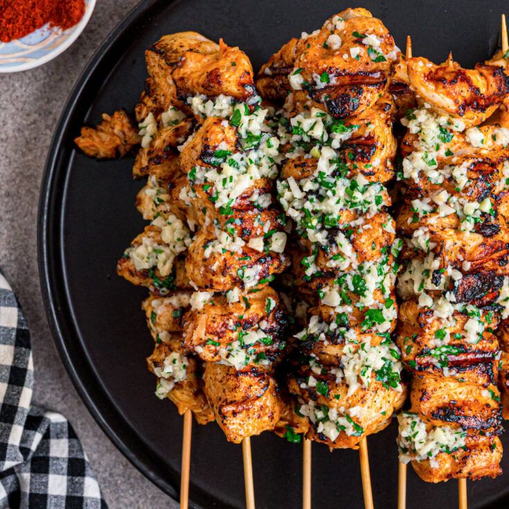 A black plate containing several perfectly grilled Garlic Parmesan Chicken Skewers loaded with parsley and garlic.