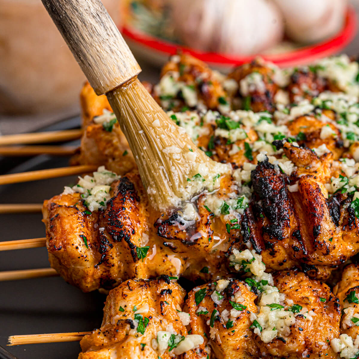 A pastry brush applies garlic butter sauce to a grilled chicken skewer.