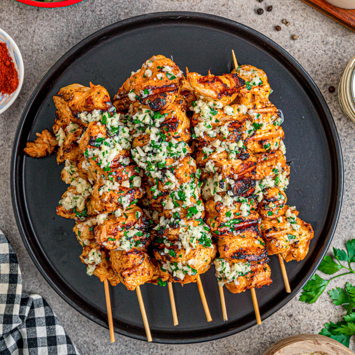 A black plate containing several perfectly grilled Garlic Parmesan Chicken Skewers loaded with parsley and garlic.