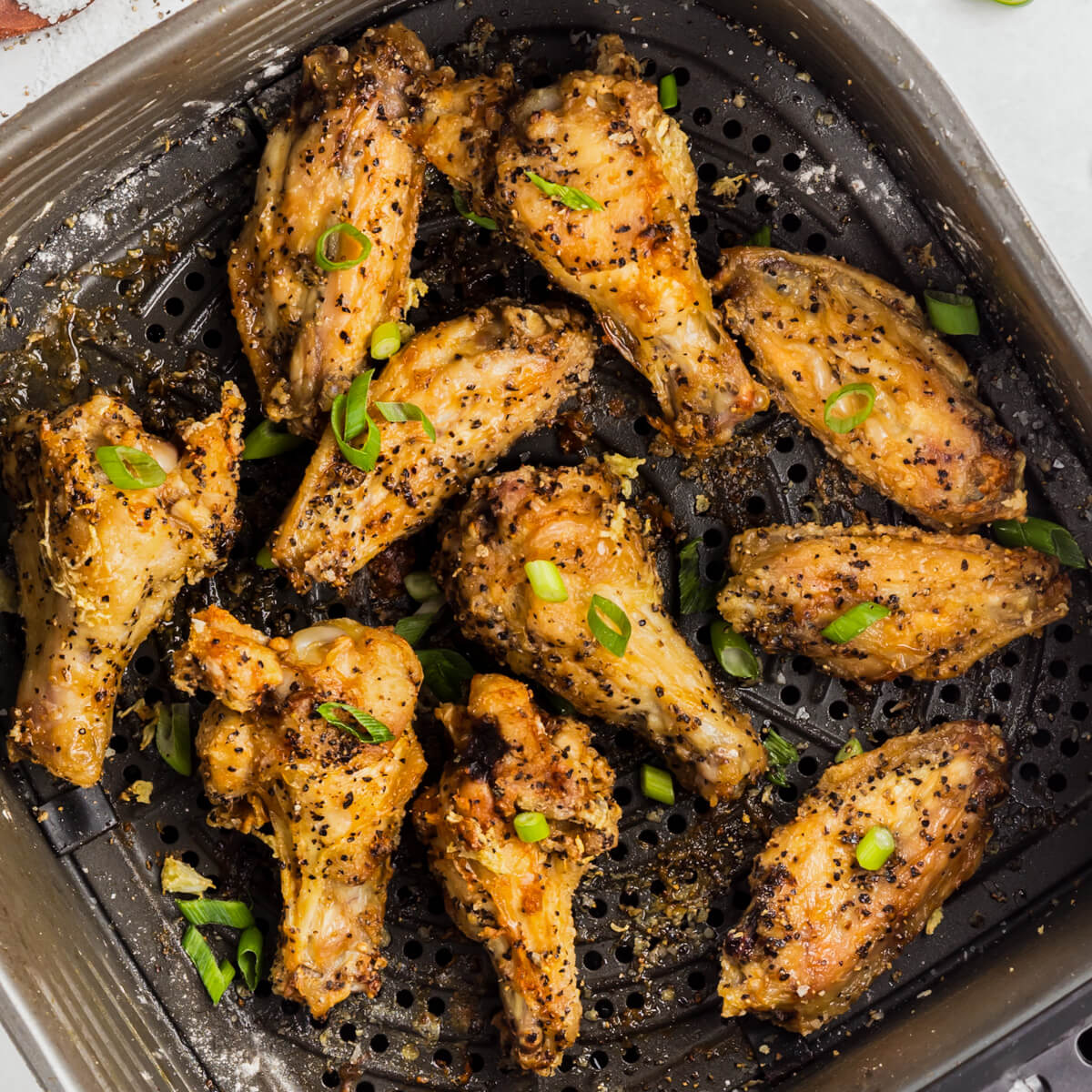 A black air fryer basket full of golden brown crispy Salt and Pepper Wings garnished with sliced green onions.
