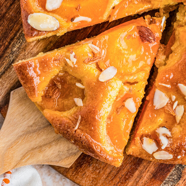 A wooden cake server holding a perfect slice of almond apricot cake dotted with baked apricots and almond slices.