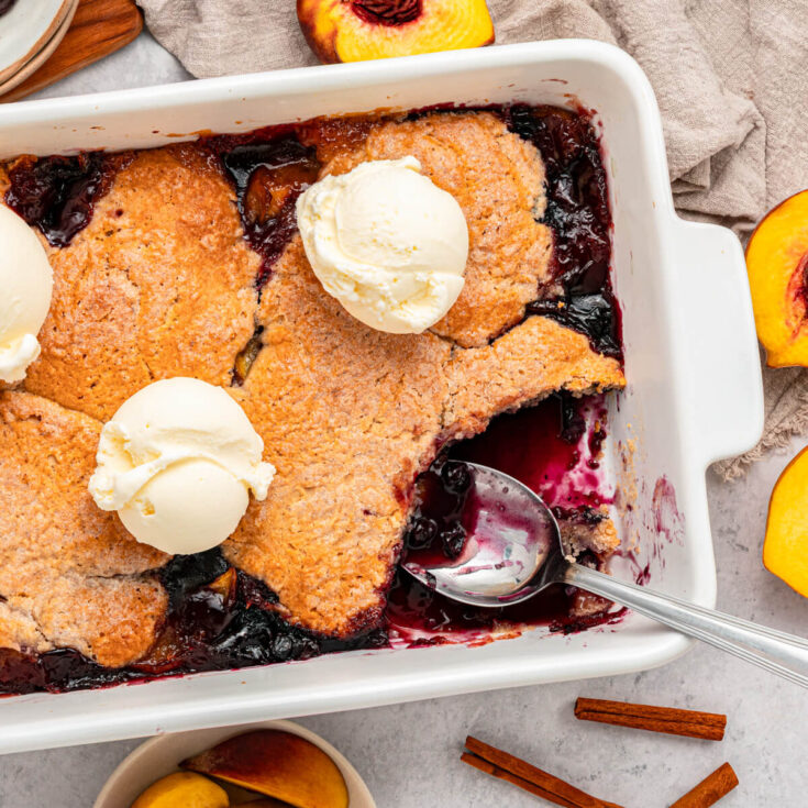 A white baking dish of Peach Blueberry cobbler topped with scoops of vanilla ice cream.