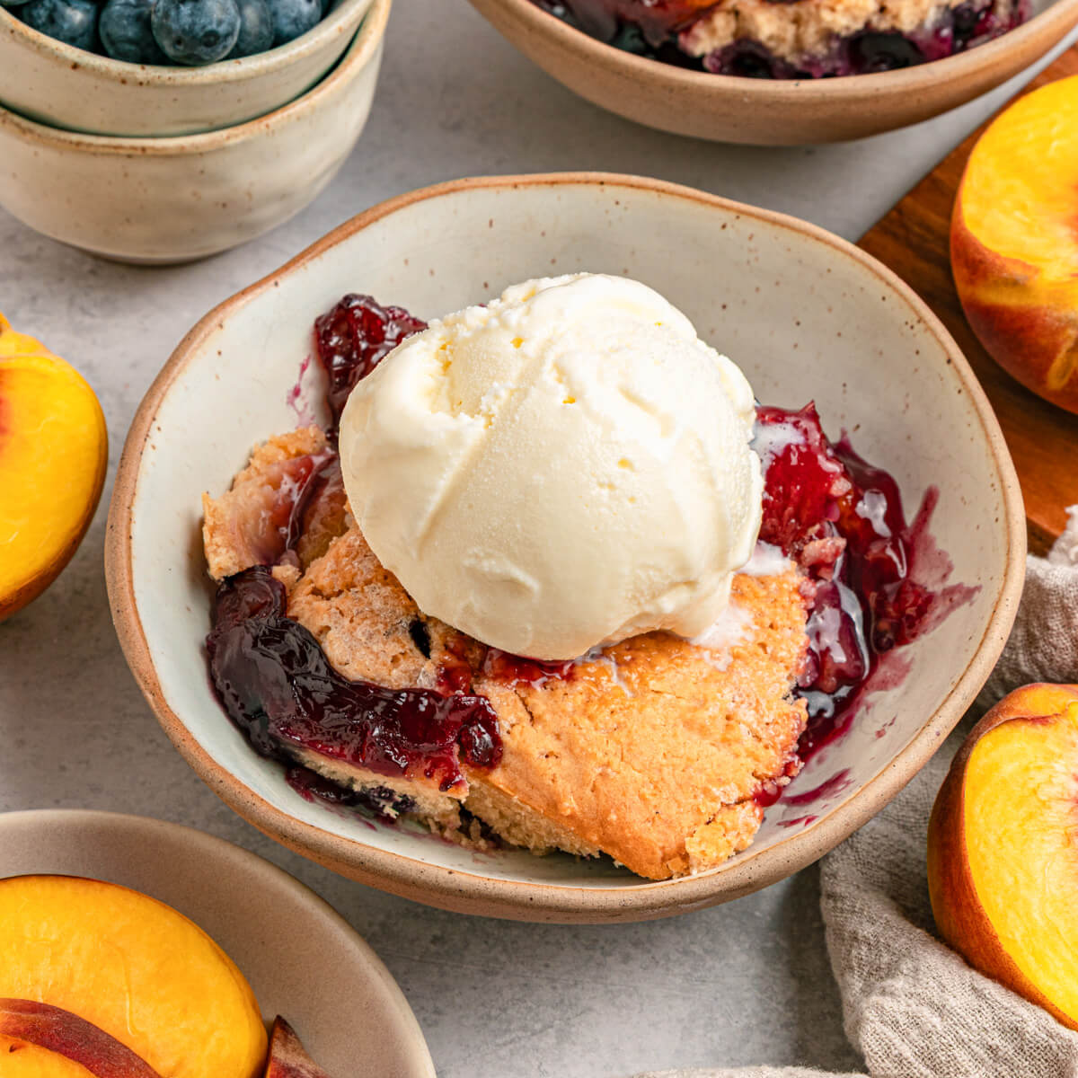 A dessert bowl of Peach Blueberry cobbler topped with scoops of vanilla ice cream.