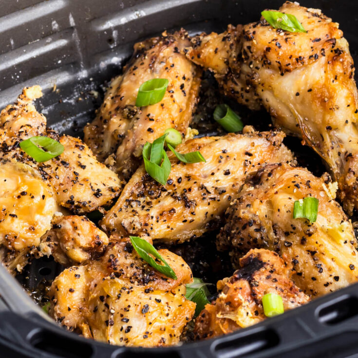A black air fryer basket full of golden brown crispy Salt and Pepper Wings garnished with sliced green onions.