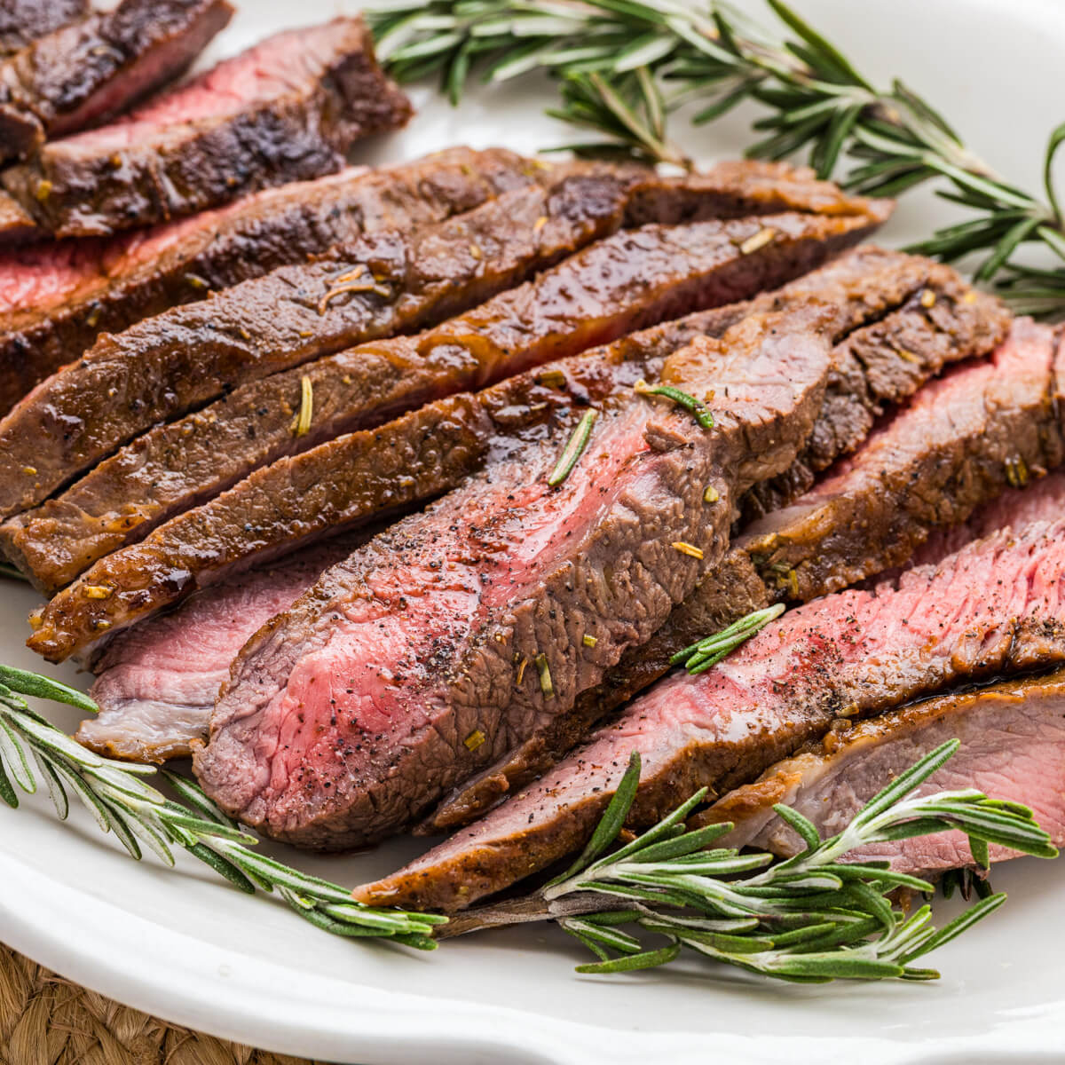 Strips of perfectly seared cast iron skillet steak surrounded by fresh rosemary on a white platter.