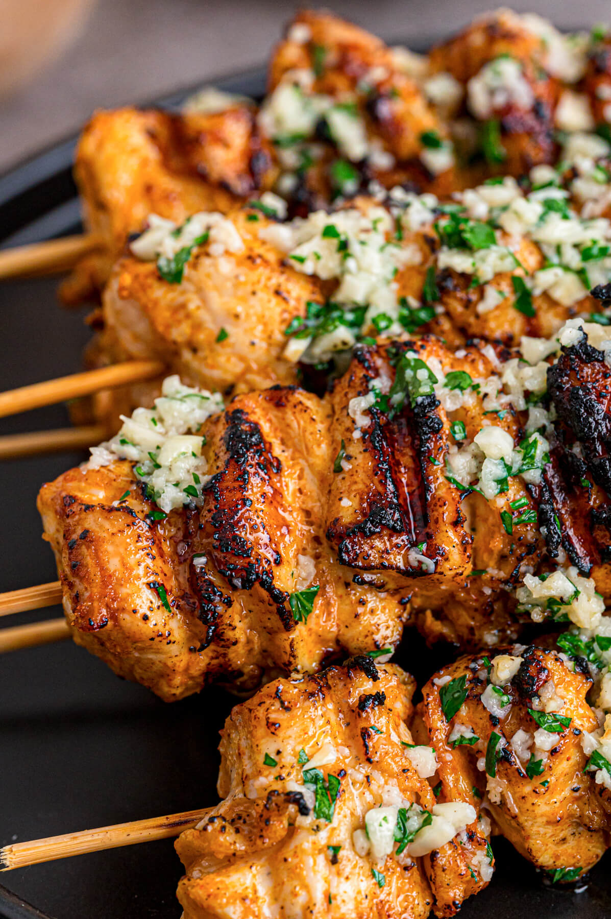 A black plate containing several perfectly grilled Garlic Parmesan Chicken Skewers loaded with parsley and garlic.