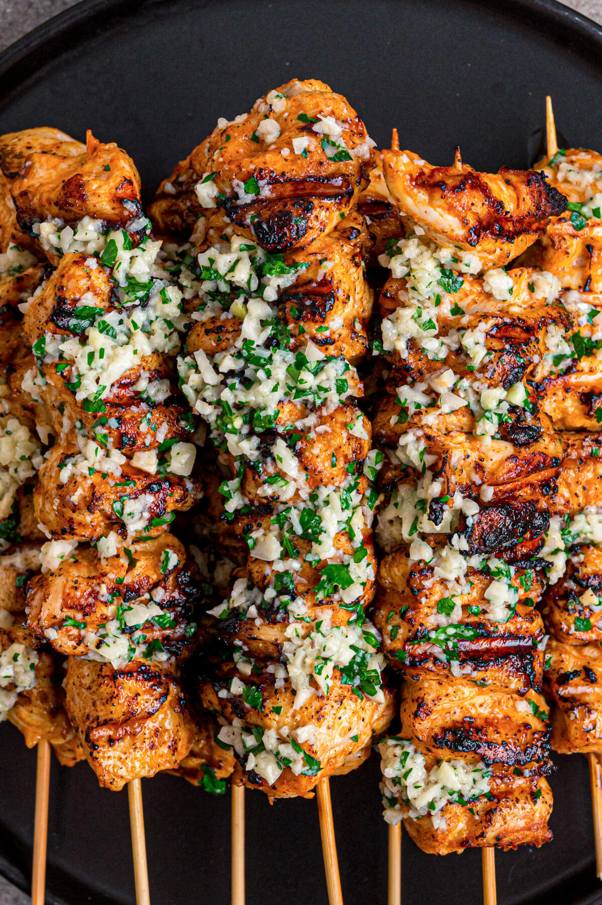 A black plate containing several perfectly grilled Garlic Parmesan Chicken Skewers loaded with parsley and garlic.