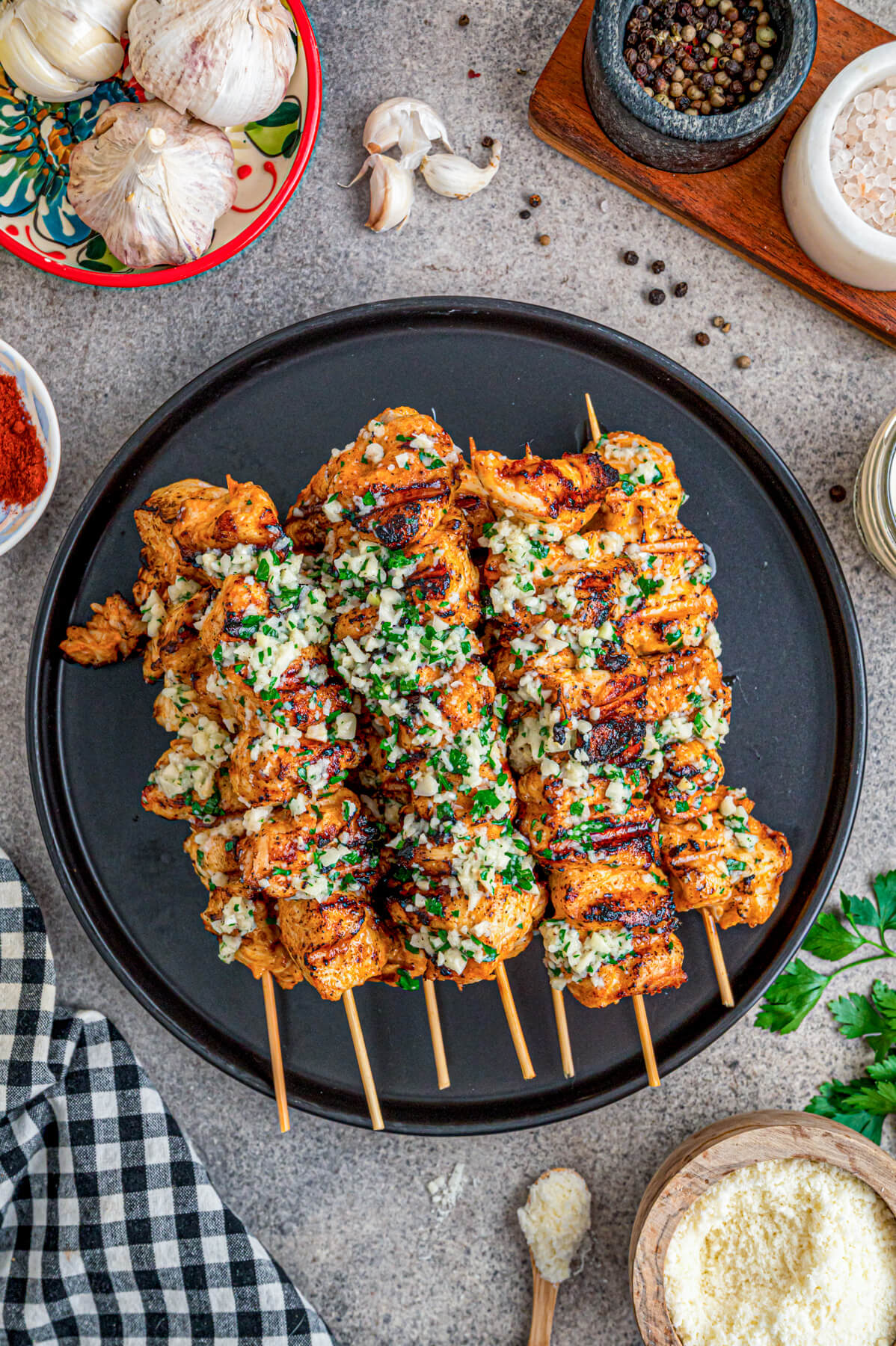 A black plate containing several perfectly grilled Garlic Parmesan Chicken Skewers loaded with parsley and garlic.