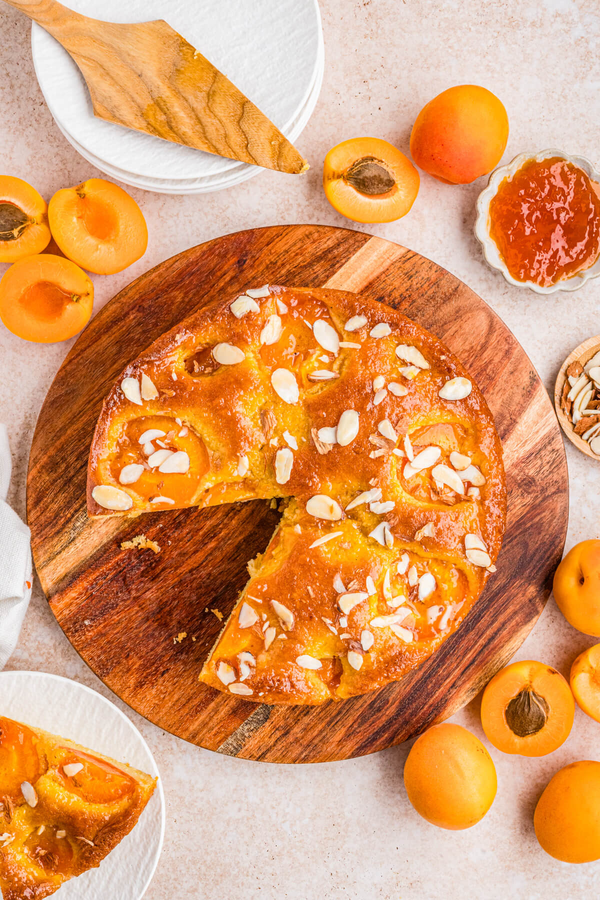 Overhead view of an almond apricot cake with one slice missing on a wooden serving board surrounded by fresh sliced apricots.