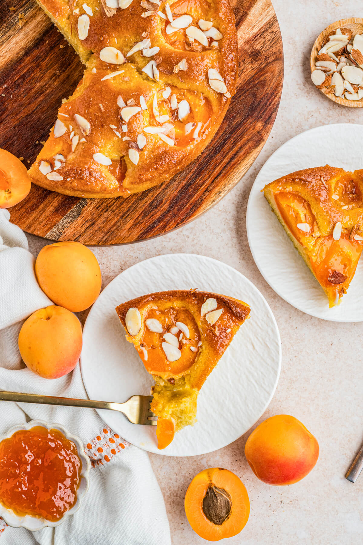 A table scene with a sliced almond apricot cake and two dessert plates holding two slices of cake among sliced fresh apricots.