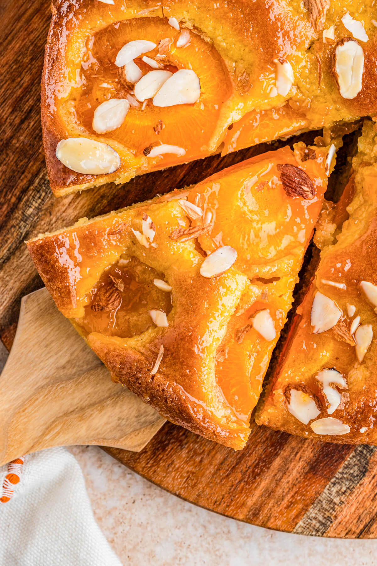 A wooden cake server holding a perfect slice of almond apricot cake dotted with baked apricots and almond slices.