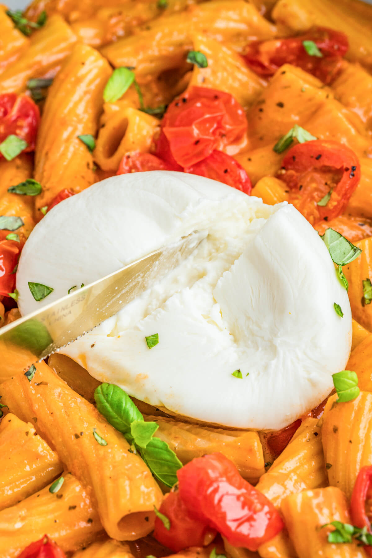 A knife slicing a burrata ball open on top of a bowl of pasta in a roasted tomato sauce.