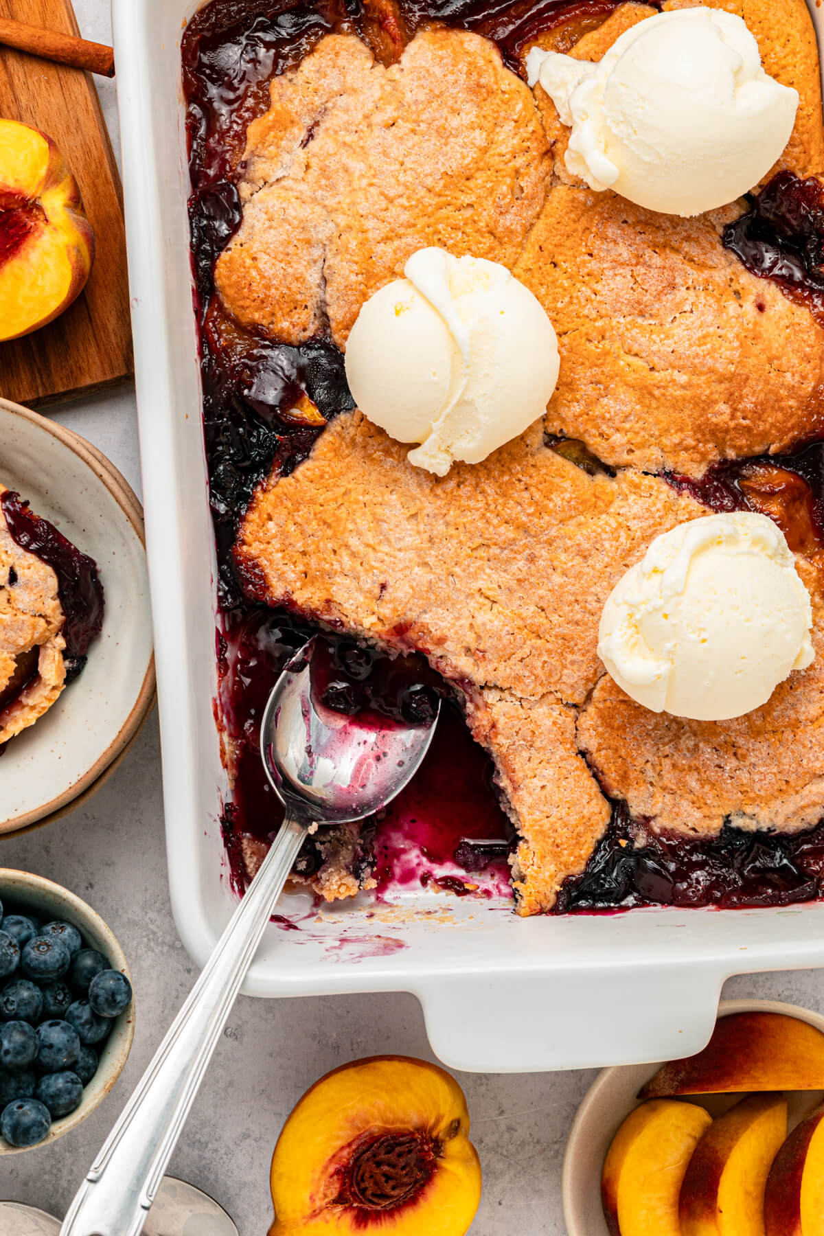 A white baking dish of Peach Blueberry cobbler topped with scoops of vanilla ice cream.