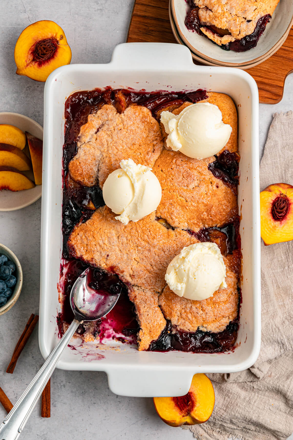 A white baking dish of Peach Blueberry cobbler topped with scoops of vanilla ice cream.