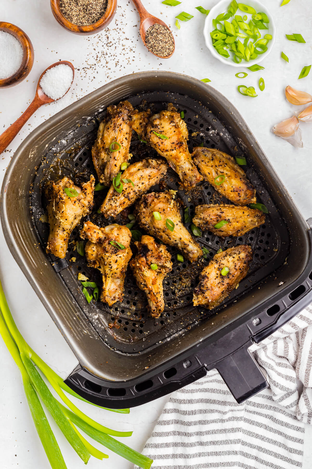 A black air fryer basket full of golden brown crispy Salt and Pepper Wings garnished with sliced green onions.