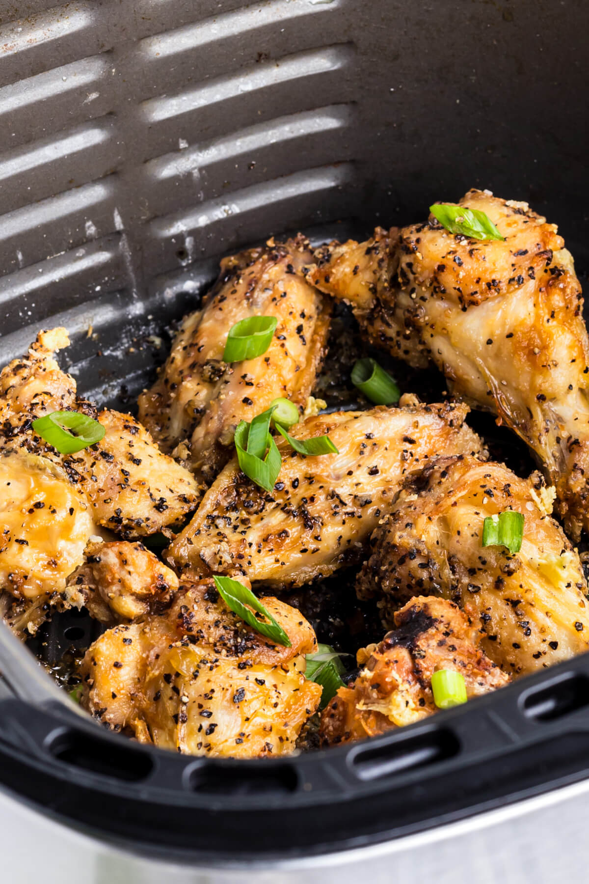A black air fryer basket full of golden brown crispy Salt and Pepper Wings garnished with sliced green onions.