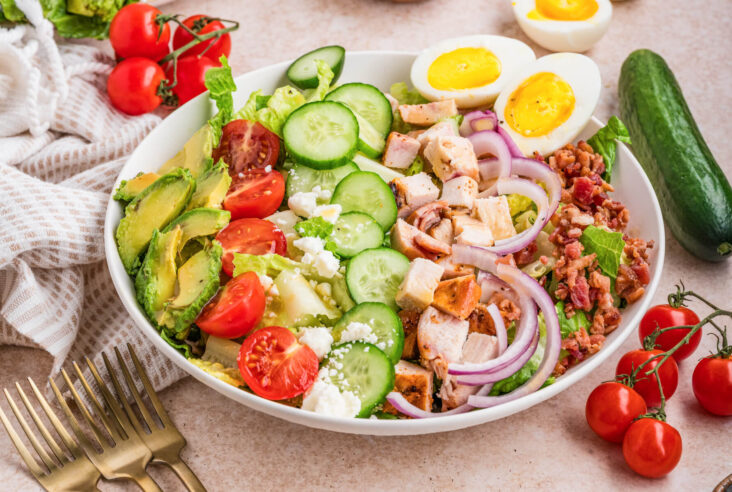 A white salad bowl containing a tastefully arranged Chicken Cobb Salad with grape tomatoes, boiled eggs, bacon, avocado, red onions, feta cheese, sliced chicken, and cucumbers.