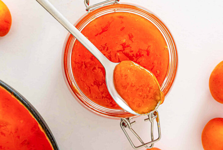 A spoon of apricot jam rests on top of a jar of the same jam surrounded by fresh apricots.