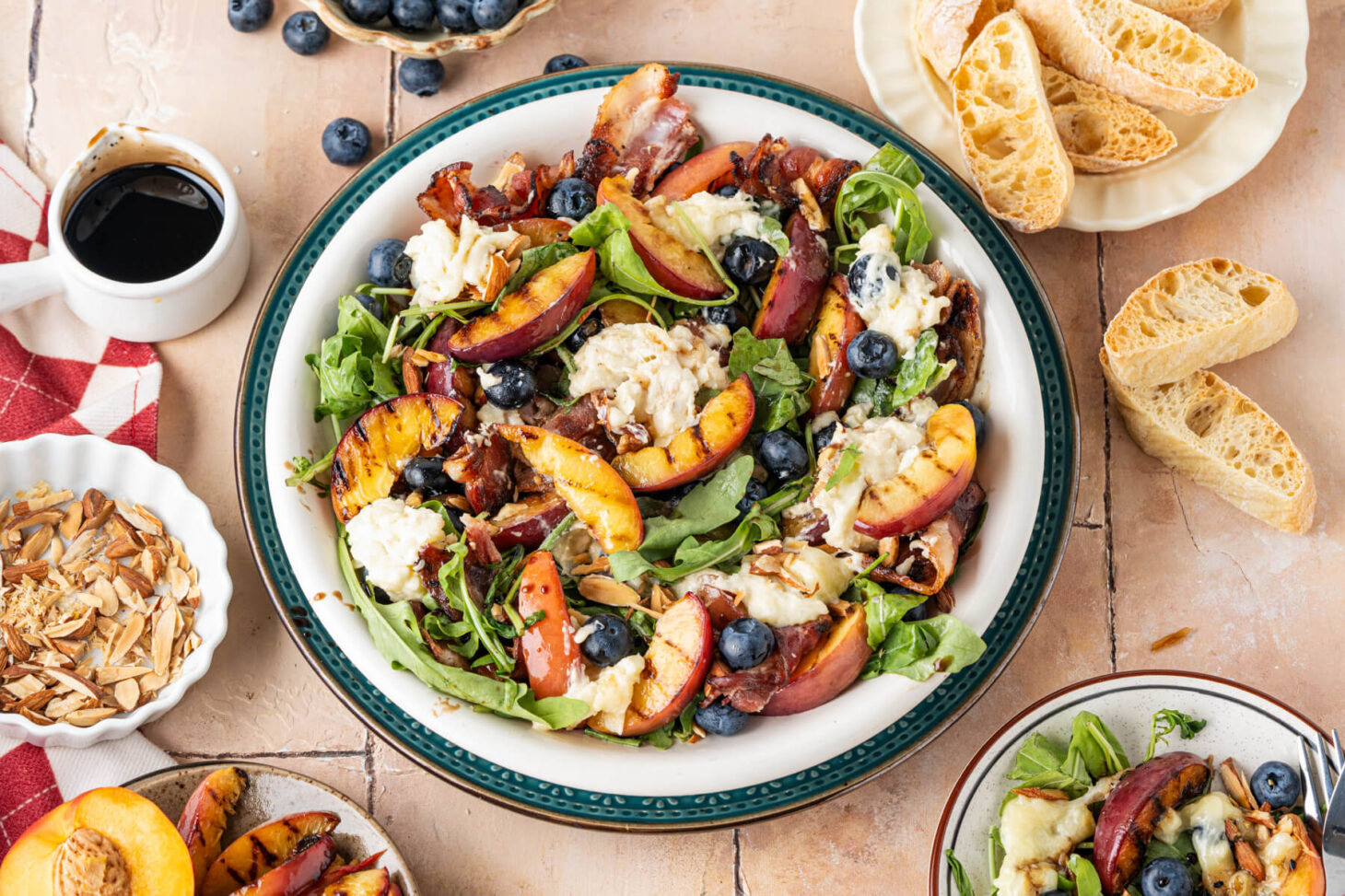 A salad bowl containing grilled peaches, toasted almonds, blueberries, crumbled bacon, and torn burrata cheese on a bed of green arugula drizzled with balsamic salad dressing.