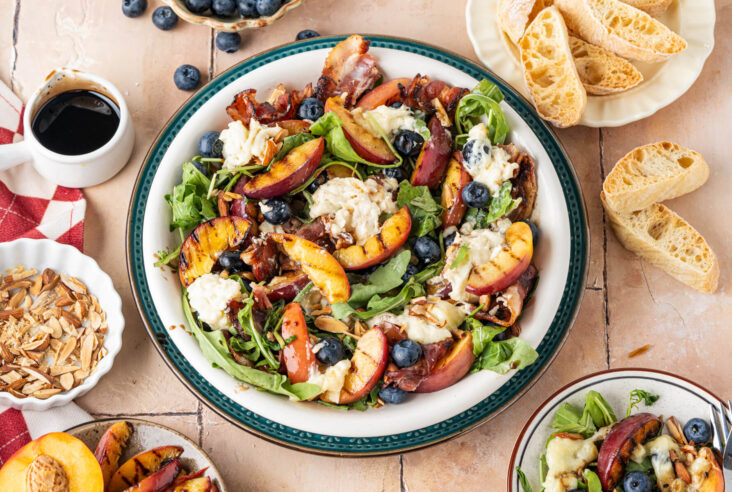 A salad bowl containing grilled peaches, toasted almonds, blueberries, crumbled bacon, and torn burrata cheese on a bed of green arugula drizzled with balsamic salad dressing.