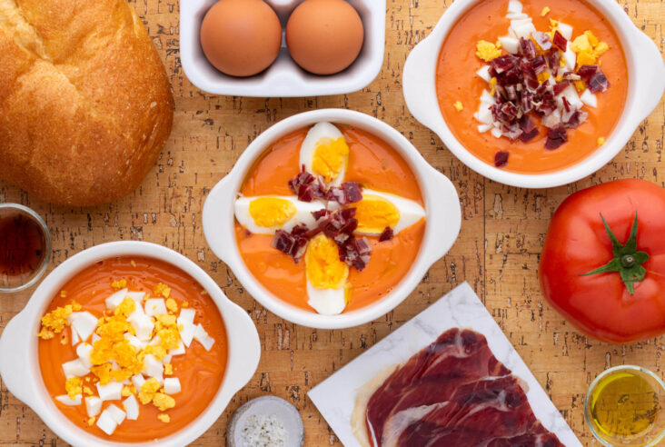 Three white bowls of vibrant orange soup garnished with chopped hard boiled eggs and jamón ibérico surrounded by recipe ingredients.