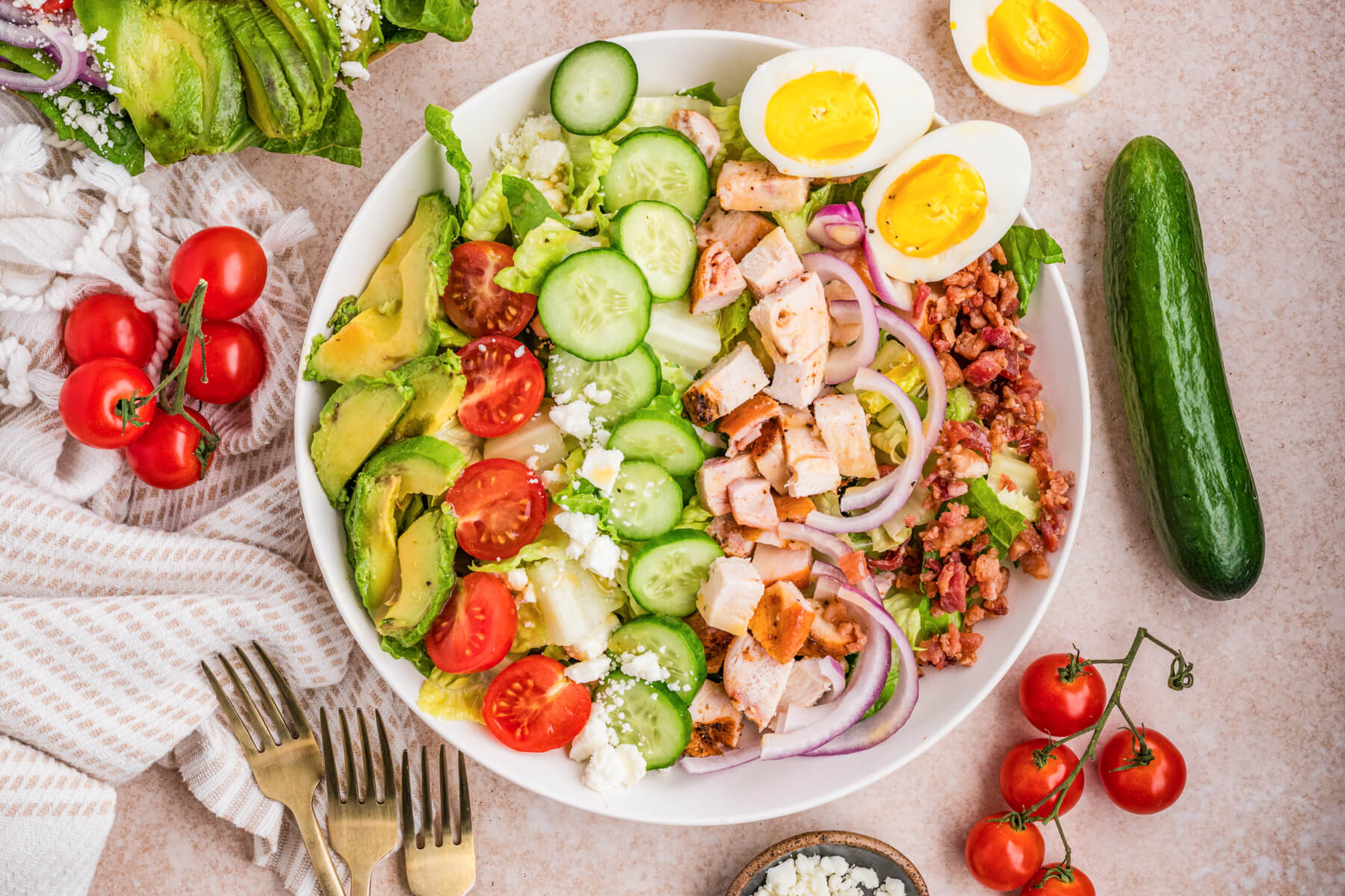 A white salad bowl containing a tastefully arranged Chicken Cobb Salad with grape tomatoes, boiled eggs, bacon, avocado, red onions, feta cheese, sliced chicken, and cucumbers.